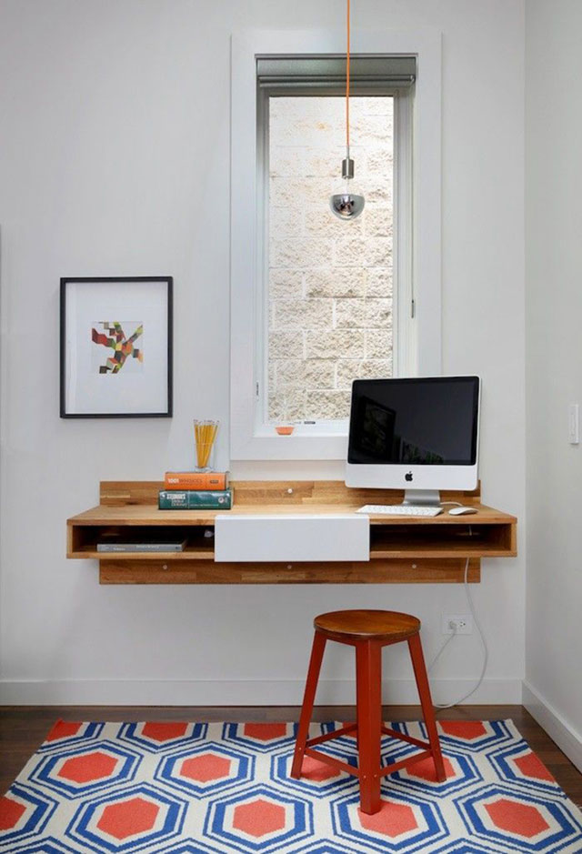 Escritorio de madera empotrado a la pared, un monitor mac, un par de libros y sobre estos un porta lápices. Piso de madera y en el muro una ilustración enmarcada.