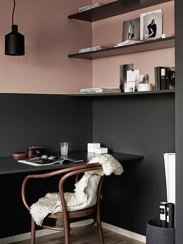 Espacio en una esquina con paredes bicolor en rosa y negro. En la mitad inferior negra una repisa voladiza también negra que sirve de escritorio, silla de madera con piel blanca como manta. Donde comienza la parte superior de color rosa, tres repisas de color negro. Lámpara negra sobre el escritorio que contrasta con el muro rosa.