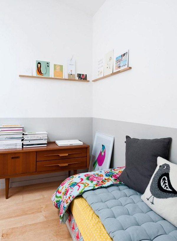 Dormitorio con diván apoyado a la pared bicolor gris y blanco. En la parte inferior gris el diván y frente a el una cómoda de madera. En la parte superior blanca solo unas repisas muy delgadas de madera con ilustraciones.