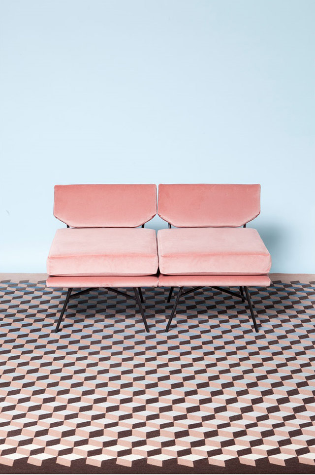 Alfombra de figura geométrica 3D en colores marrón claro y oscuro con azul pastel. Sofá dos cuerpos en tela color rosa y patas delgadas metálicas. Pared posterior en celeste.