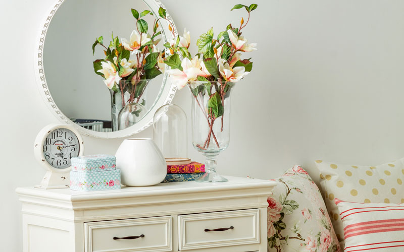 Cómoda blanca con cajonera, adornada con un reloj y florero, mostrando un espejo y parte de un sillón con cojibes de distintos diseños.