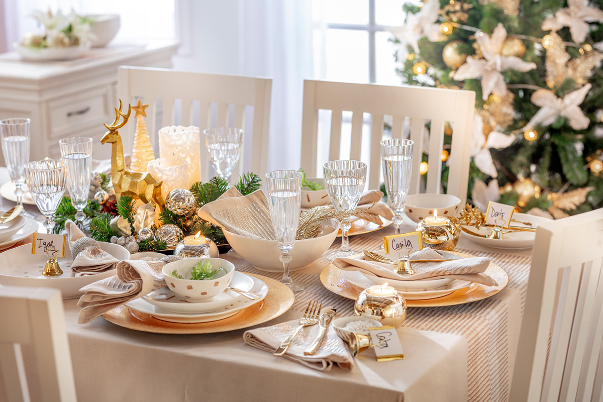 Vista general de mesa decorada con adornos y tonos navideños. Mantel color crema con detalles dorados, velas y blanco de fondo.