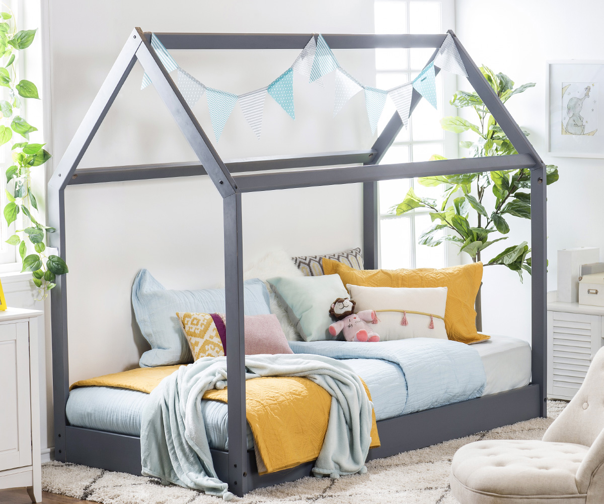 Vista general de dormitorio infantil. Se puede ver una cama de madera que tiene estructura de casa. Va directamente sobre una estructura de madera pegada al suelo y sus mantas celestes y mostaza le dan un toque relajado pero alegre.