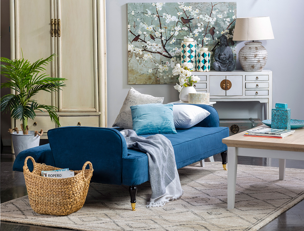 Vista general de sala de estar con futón extendido como cama, con cojines y manta. Mesa de centro de base blanca con superficie color madera.